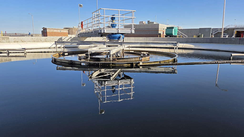 Circular clarifier tank at a municipal water treatment plant, with a central rotating mechanism and walkway, reflecting the clear blue sky on the water's surface.