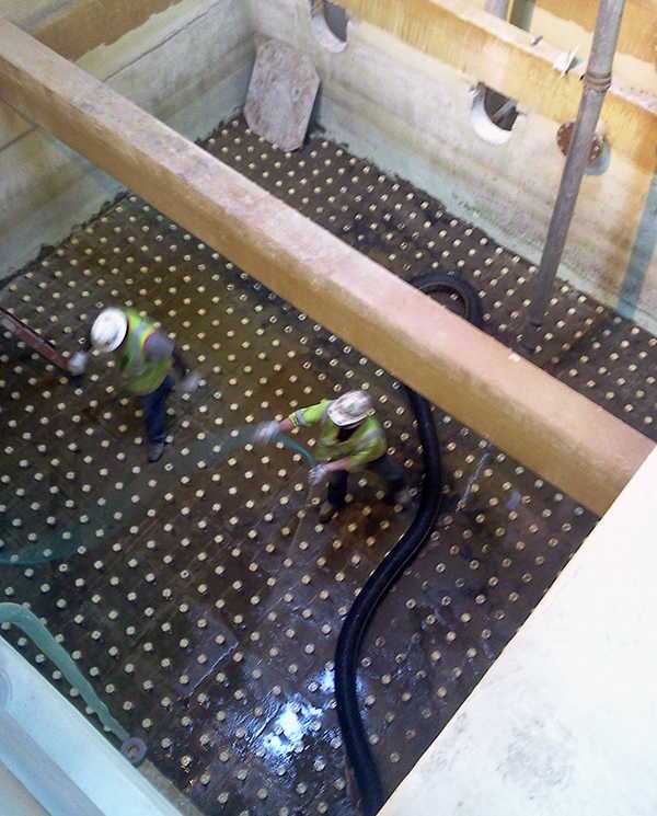 A downward view into the bottom of a rectangular concrete filter cell showing two plant workers, one is spraying the floor of the filter underdrain with a hose.