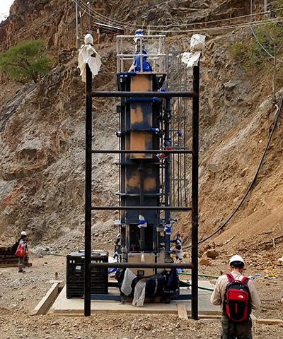Tall, narrow, cylindrical pilot clarifier resting on the canyon floor below the mine, a caged area and drive unit sit atop the clarifier.