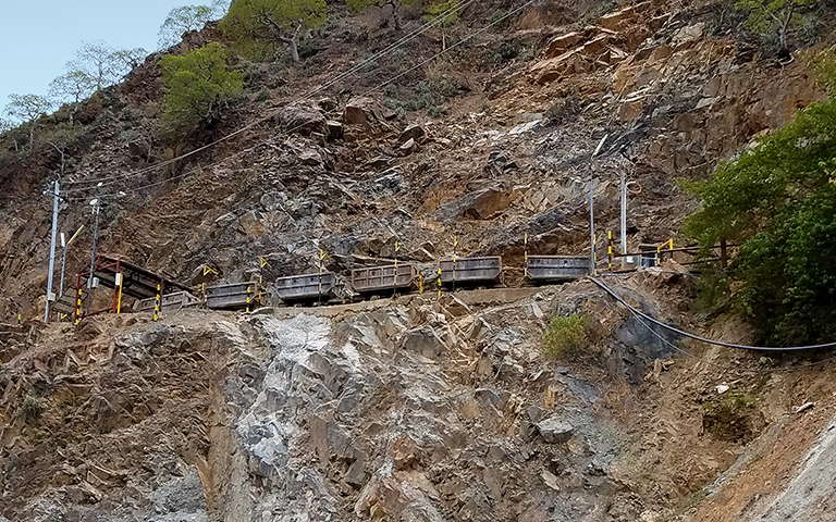 Seven rail cars running horizontally along a ledge cut into a steep hillside with exposed rock at the mine site